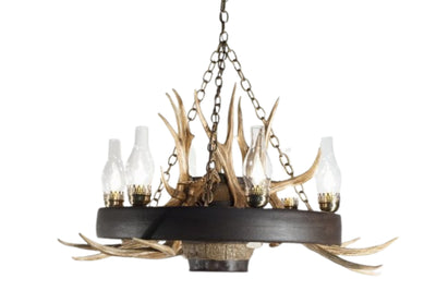 A rustic wagon wheel chandelier with mule deer antlers and chimney glass covering the lights.