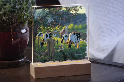 Acrylic table top night light featuring cows grazing on a country farm with a wood base sitting on a table with a potted plant in the background.