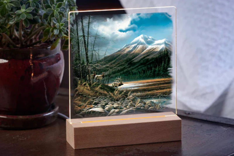 Acrylic table top night light featuring three elk looking over a high mountain meadow with a snowy peak in the background sitting on a table with a potted plant in the background.