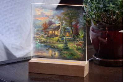 Acrylic table top night light featuring the artwork by Thomas Kinkade.  Sitting on a table top with a potted plant in the background.