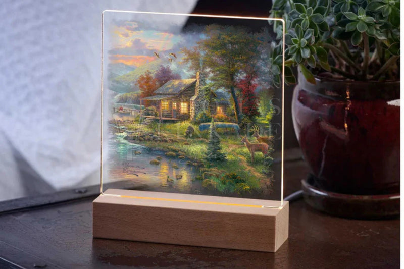 Acrylic table top night light featuring the artwork by Thomas Kinkade.  Sitting on a table top with a potted plant in the background.