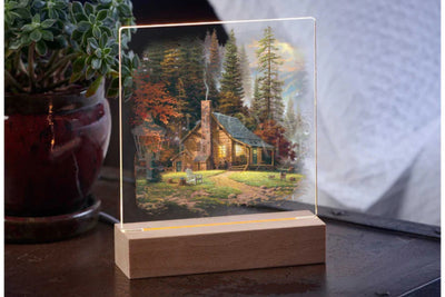Acrylic tabletop night light featuring a peaceful secluded cabin in the woods with a wood base sitting on a table with a potted plant in the background.