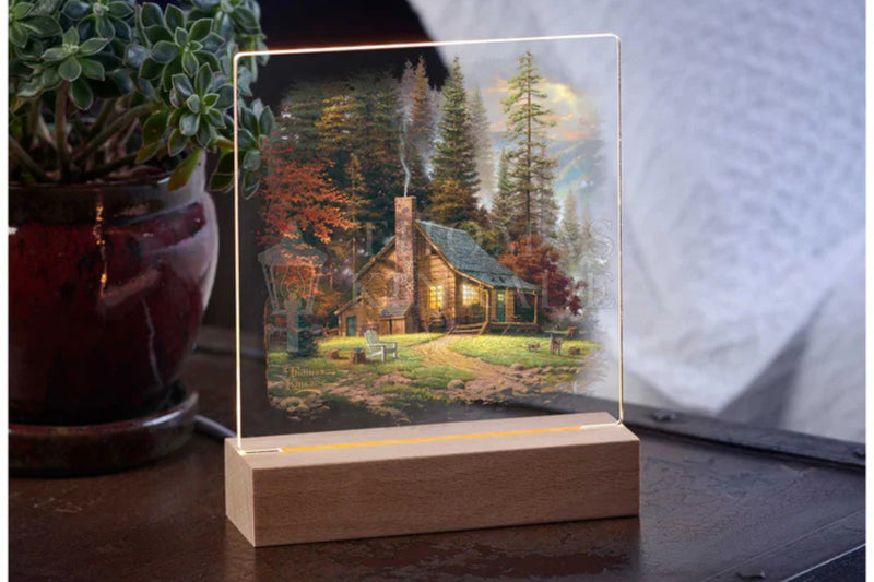 Acrylic tabletop night light featuring a peaceful secluded cabin in the woods with a wood base sitting on a table with a potted plant in the background.