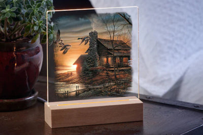 Table top acrylic night light featuring scenic design of a lakeside lodge with flying waterfowl and a distant sunset on a wooden base.  The light is on a table with a potted plant in the background.