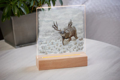 Mule Deer Design Acrylic tabletop night light with a wood base sitting on a table with a potted plant in the background.