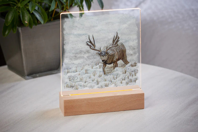 Mule Deer Design Acrylic tabletop night light with a wood base sitting on a table with a potted plant in the background.