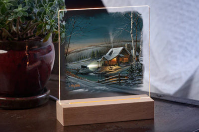 LED tabletop night light featuring a scenic picture of an old car approaching a snowy cabin in the woods sitting on a wood base with a potted plant in the background.
