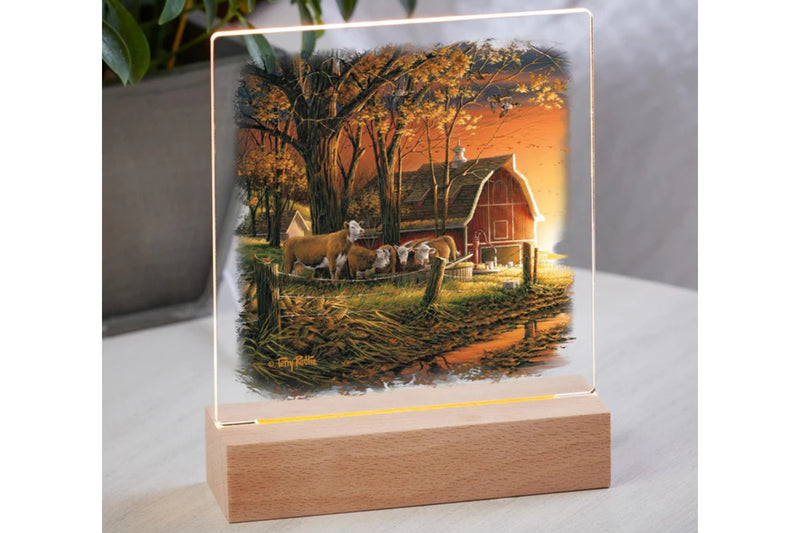 Acrylic barn with a cow design tabletop night light with wood base sitting on a table with potted plant in the background.