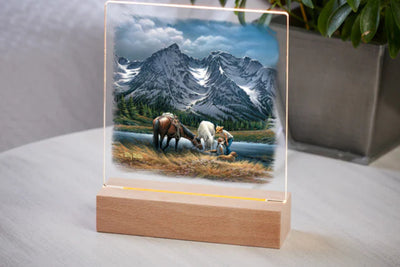 Acrylic night light featuring an image of a couple, their two horses and companion dog with a snowy mountain in the background on a wooden base sitting on a table with a potted plant in the background.
