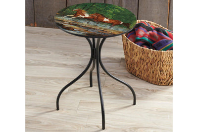 Small round metal side table with a painted design of horses running through a mountain creek on a wooden floor, next to a woven basket with a blanket.